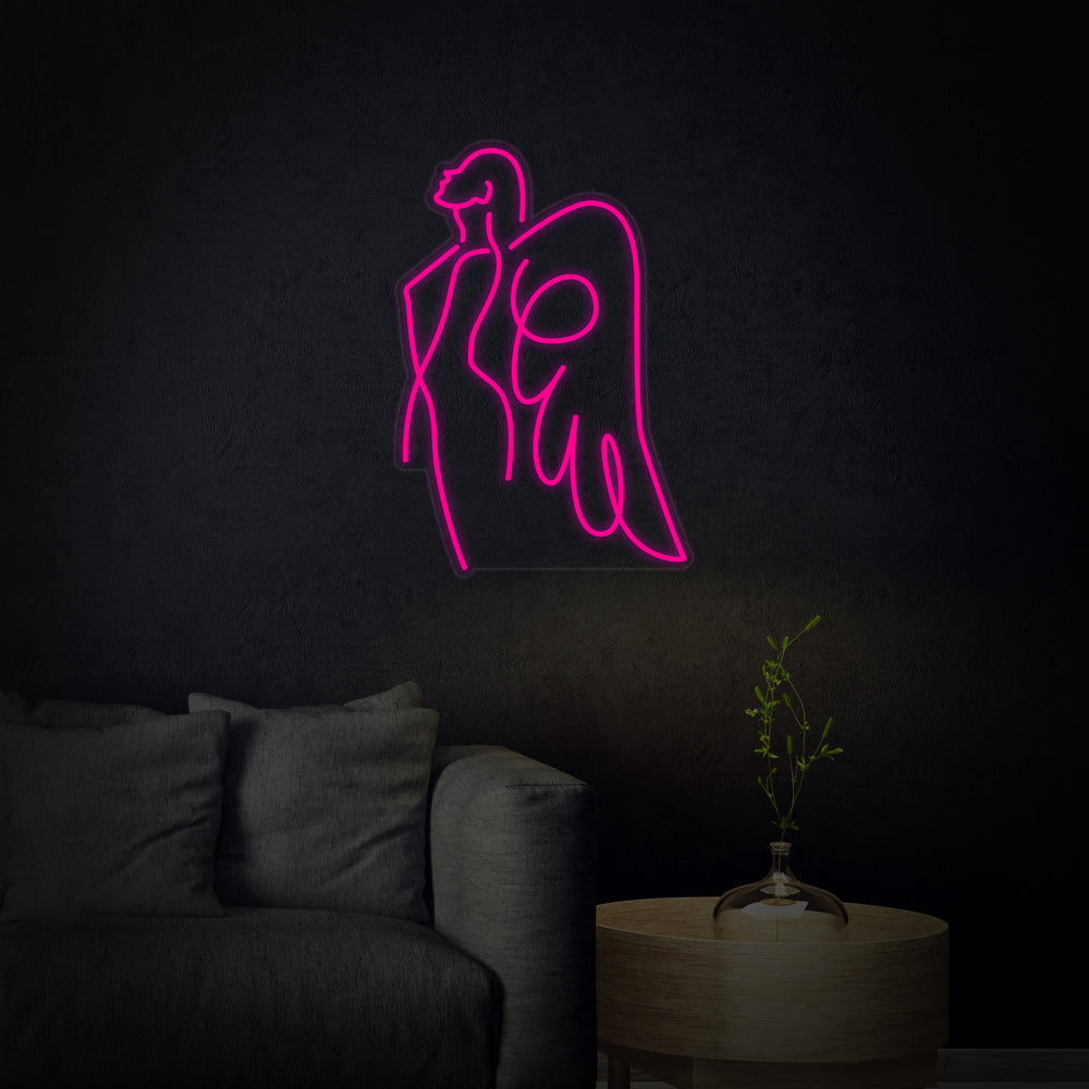 Artistic Angel Mom script neon sign glowing with soft white light on a dark, reflective wall with subtle wings graphic.
