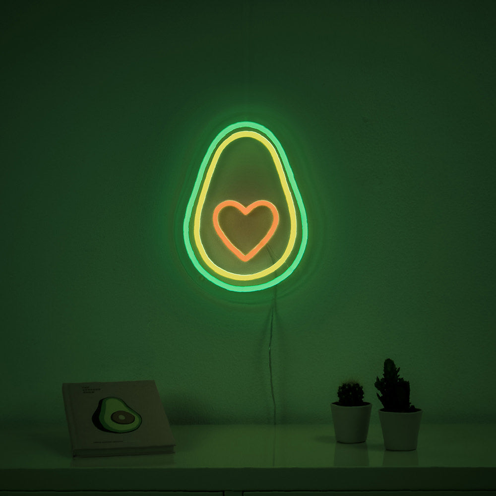 A real neon sign of an avocado with a heart shape inside, showing physical tubing, wires, and clear acrylic backing, resting on a wooden surface.