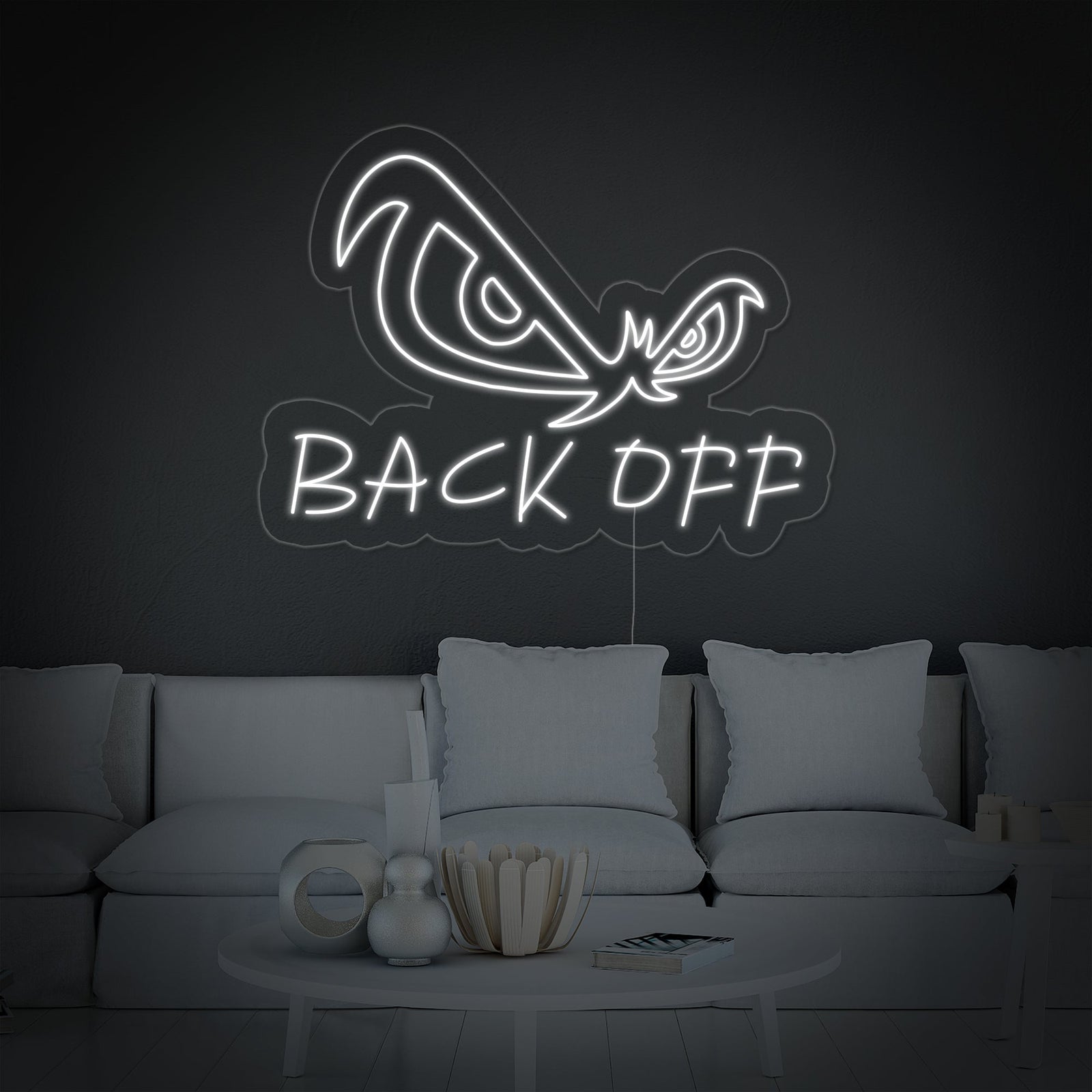 A vibrant 'Back Off Eagle Eyes' neon sign glows blue in a contemporary living room setting, mounted on a grey wall above a sleek black sofa with patterned cushions.