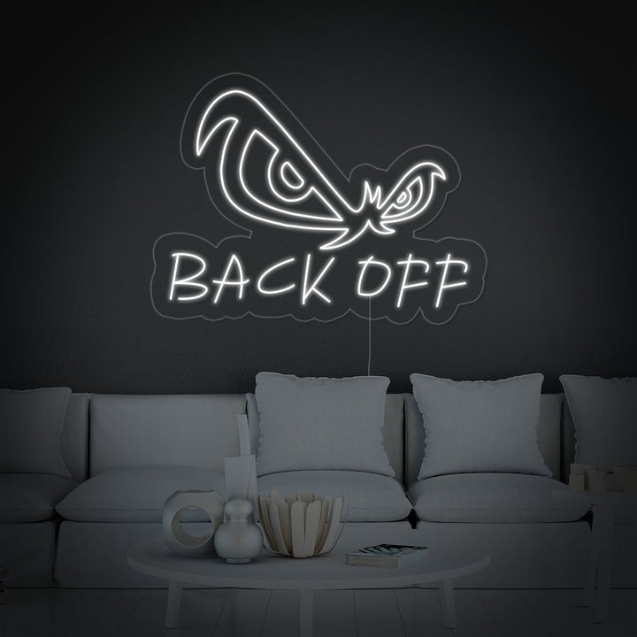 A vibrant 'Back Off Eagle Eyes' neon sign glows blue in a contemporary living room setting, mounted on a grey wall above a sleek black sofa with patterned cushions.