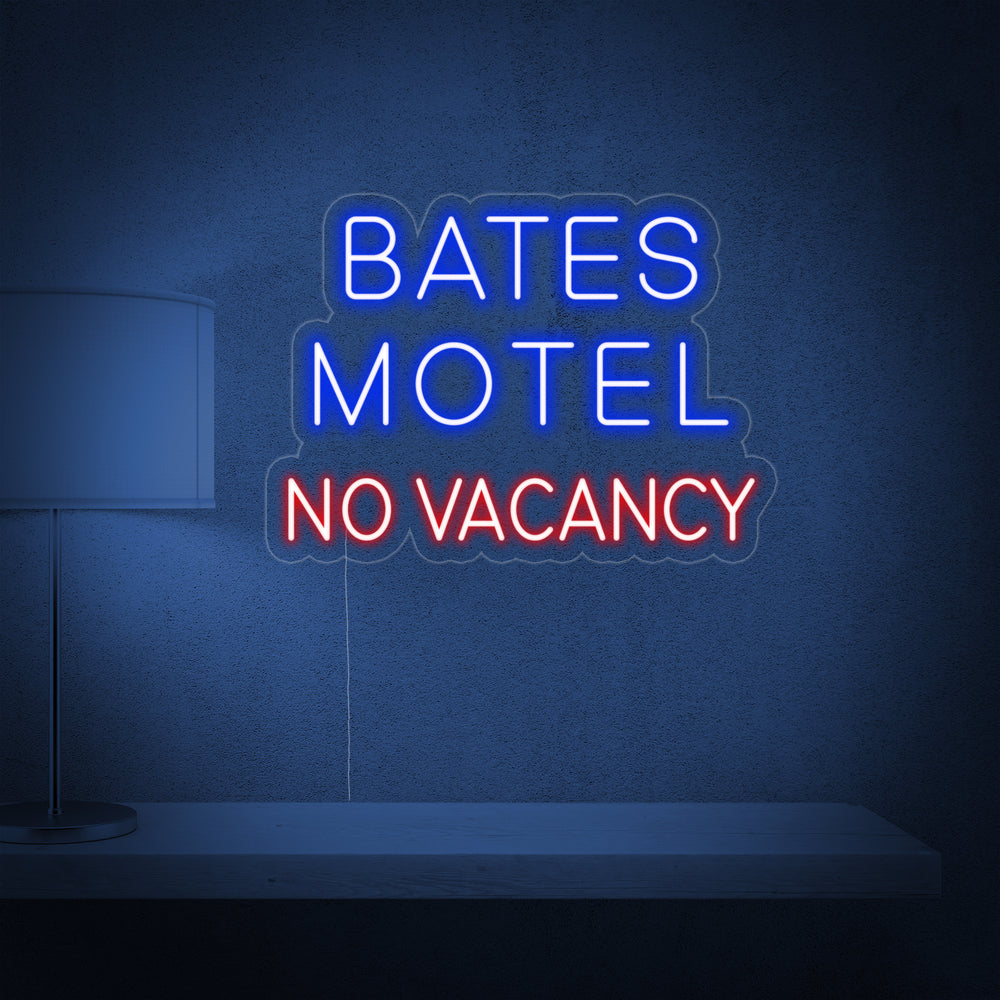 A glowing red and white neon sign displaying 'Bates Motel' and 'No Vacancy' mounted on a dark brick wall above a vintage lamp and shelf.