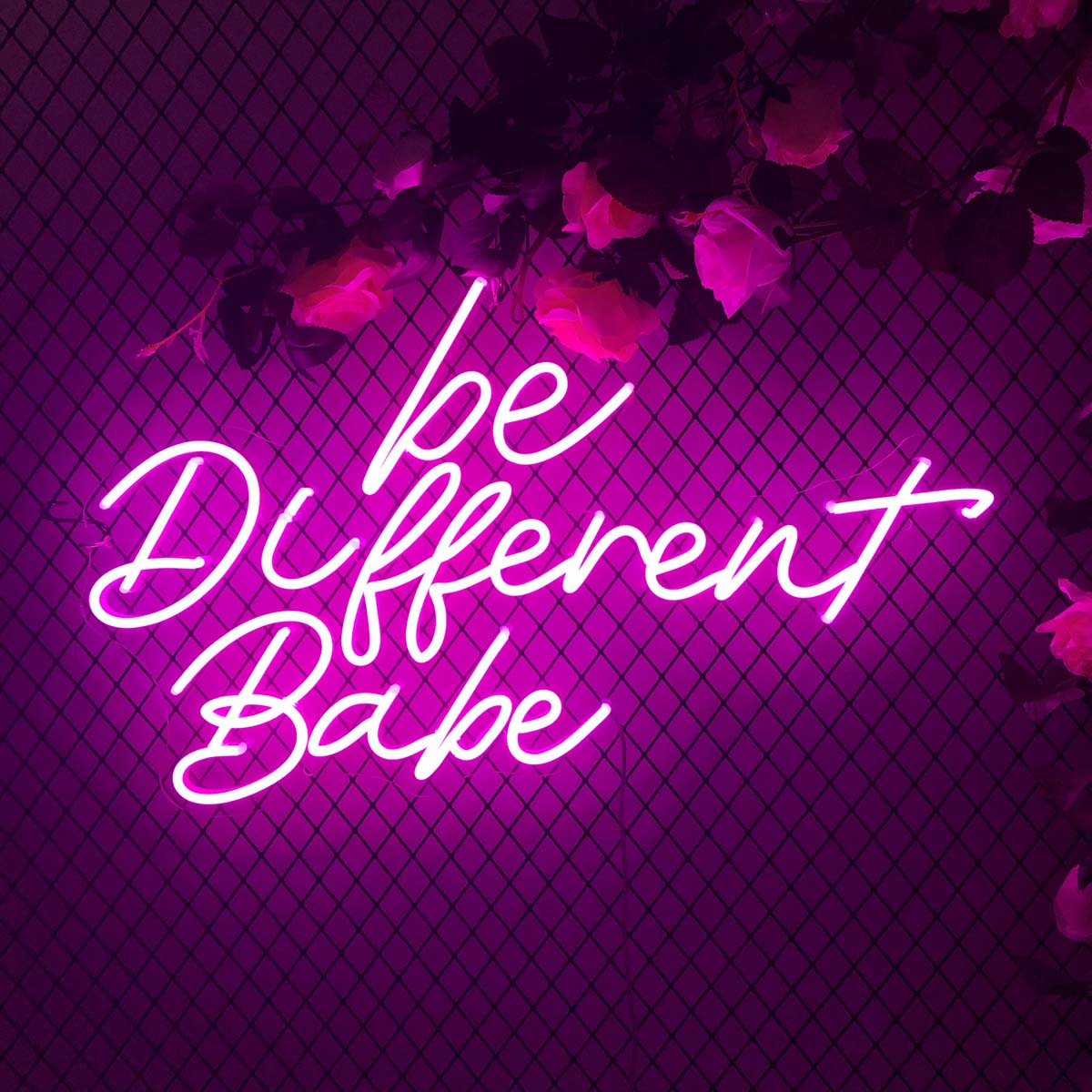 A vibrant pink "Be Different Babe" neon sign in stylized script, glowing brightly on a black wire mesh wall decorated with flowers.