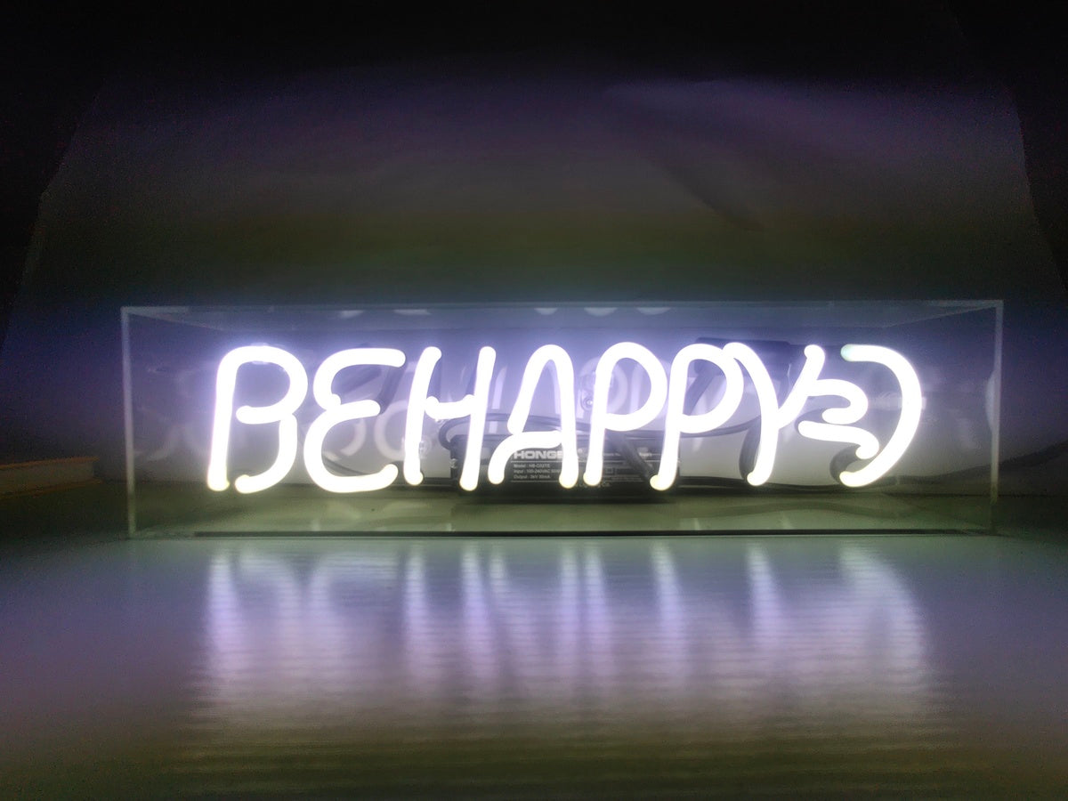 A rectangular clear acrylic box containing a hand-bent neon sign illuminating the words 'Be Happy' in blue, with a visible power cord and transformer sitting on a reflective surface.