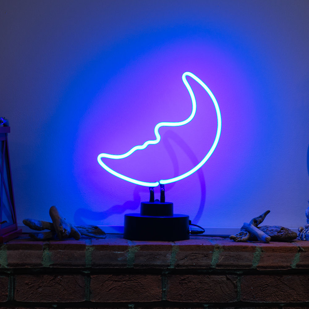 A lit Blue Moon neon sign displaying the brand logo with moon and text, sitting on a wooden desk next to a blurred plant and brick wall.