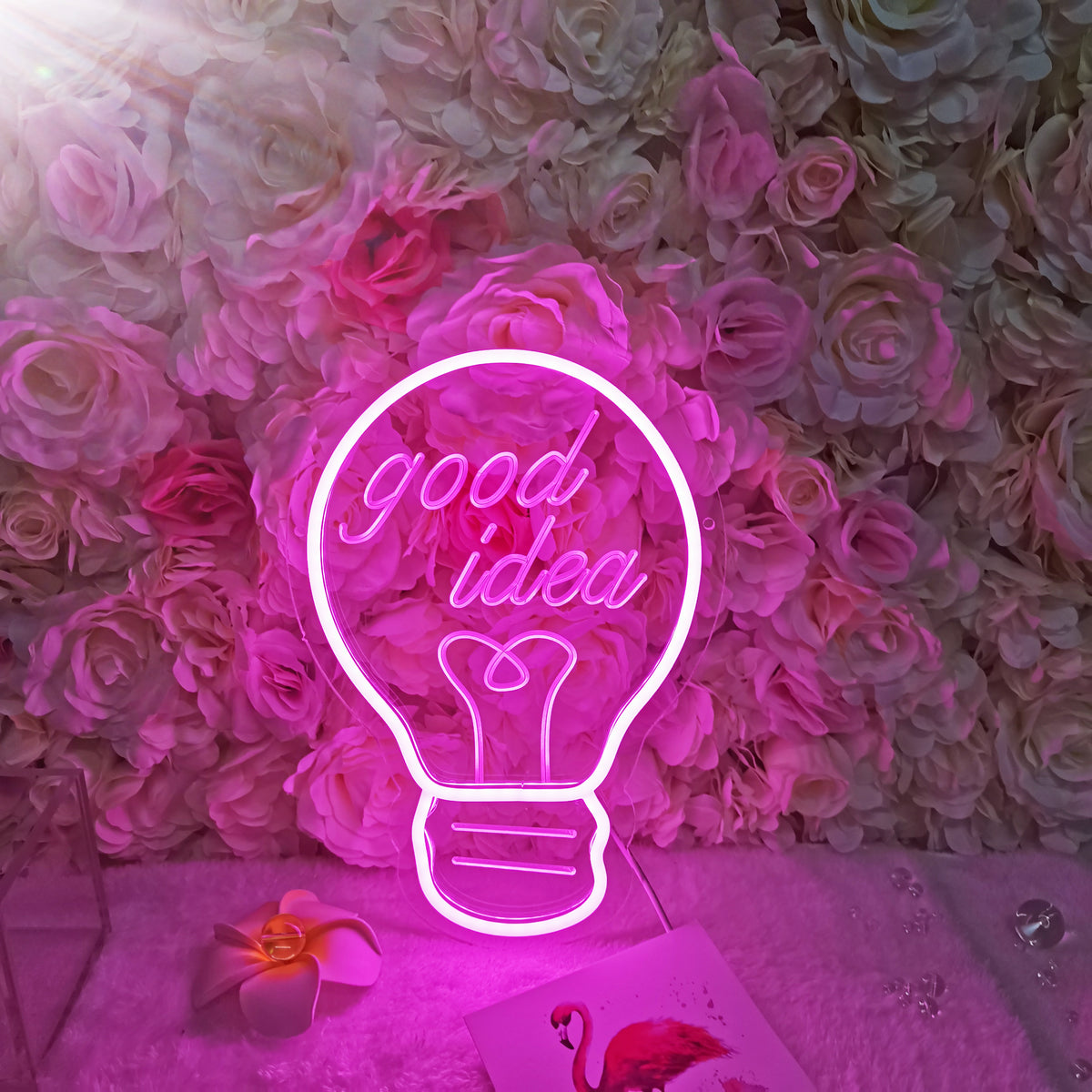 Creative 'Bulb Good Idea' dimmable neon wall decor above a light pink desk, next to potted plants and a fuzzy chair.