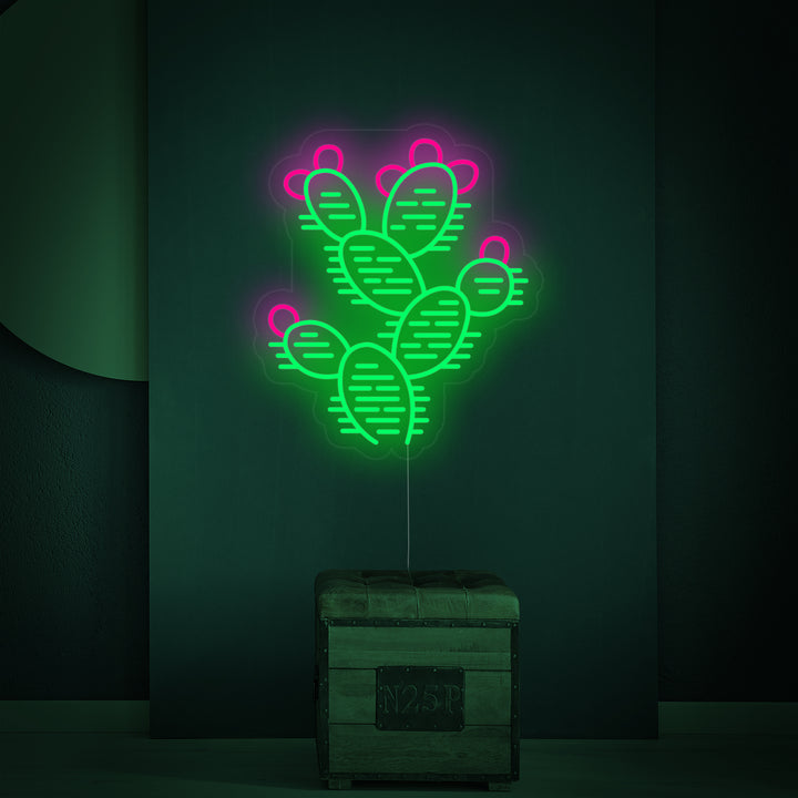 A stylized green cactus neon sign on a rectangular clear backing, mounted on a plain grey wall with the light on.