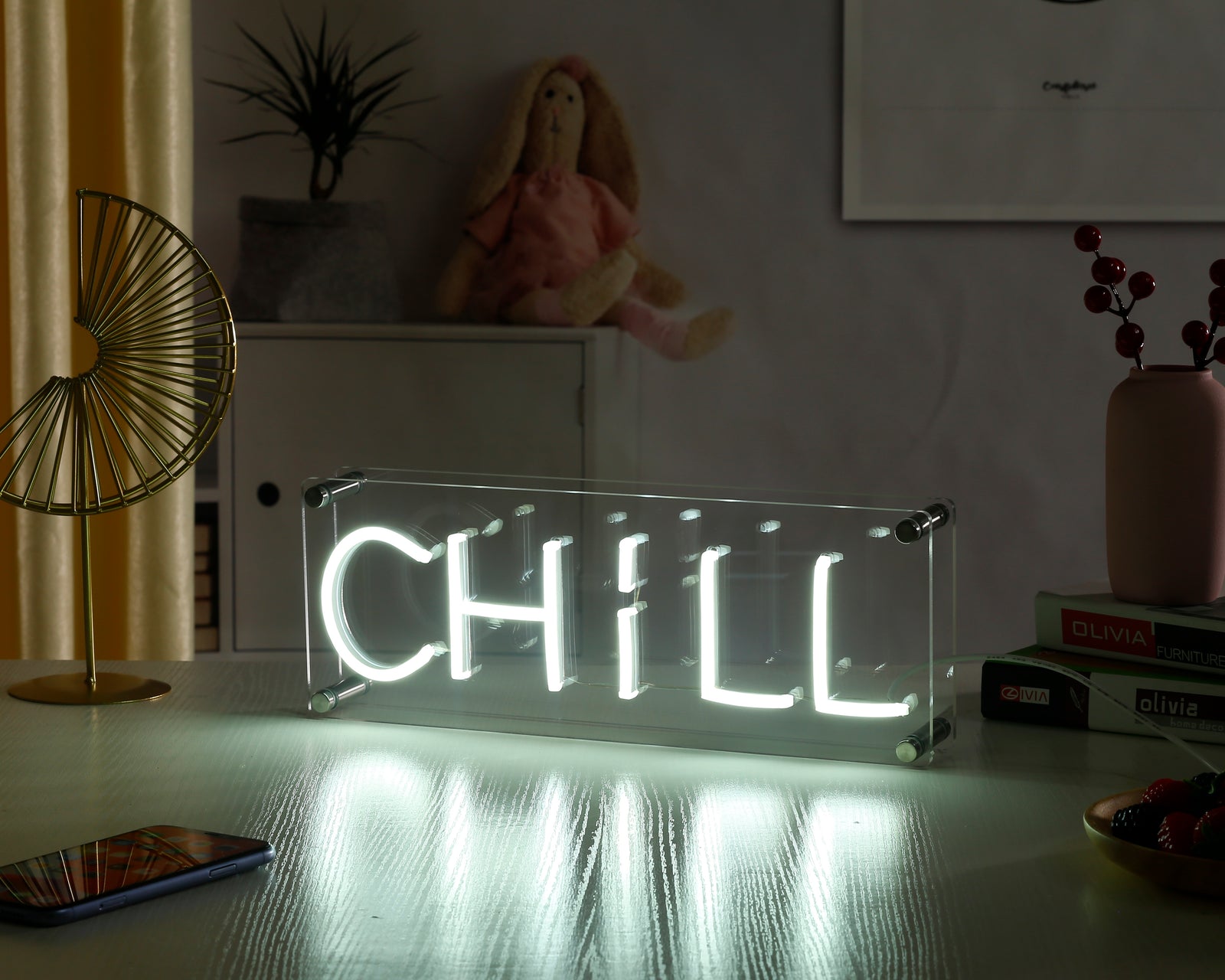 A 'Chill' neon desk sign with blue letters glowing on a clear acrylic stand placed on a wooden desk near potted plants.
