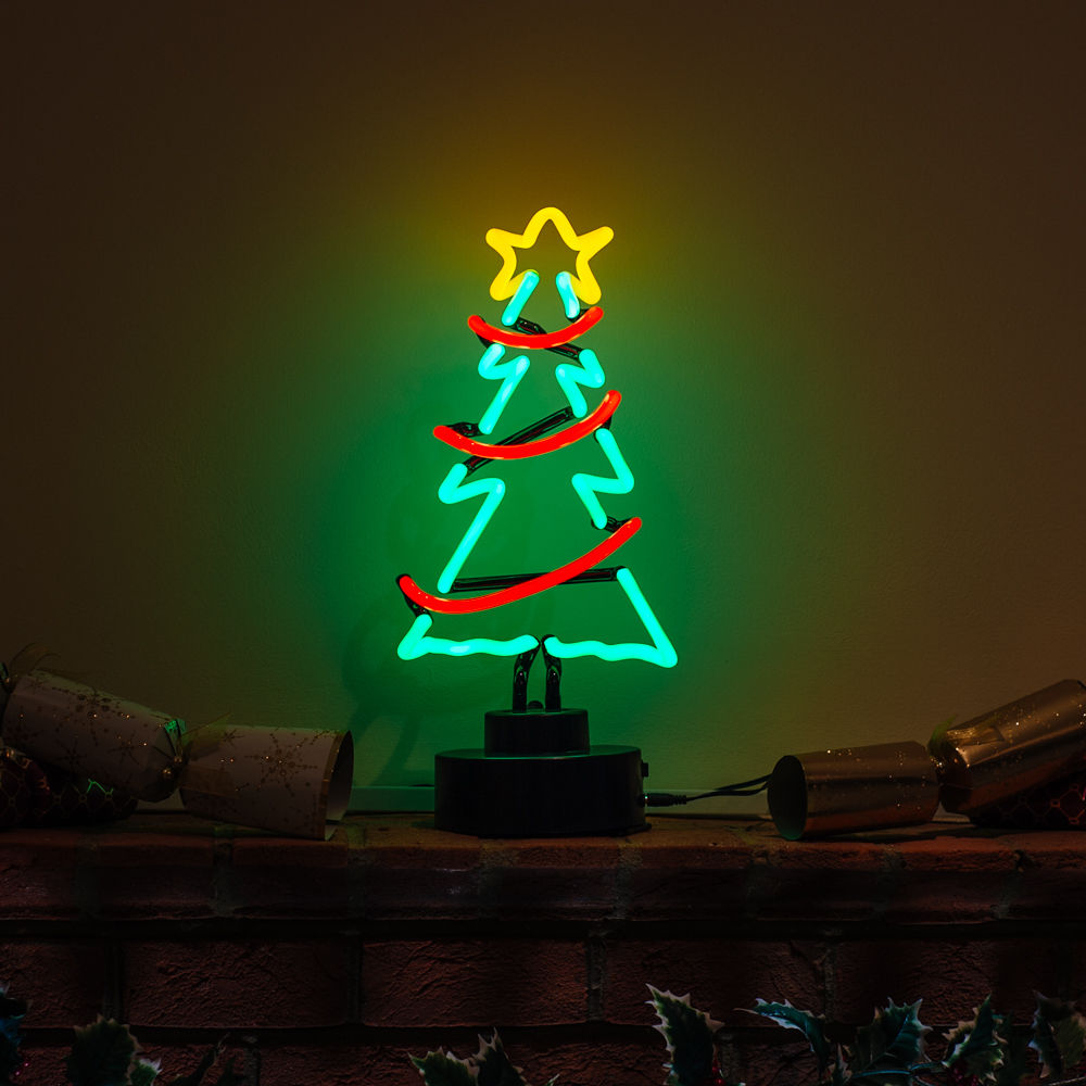 A glowing green 'Christmas Tree' neon sign on a simple black base, illuminating a festive scene with Christmas crackers and holly leaves on a table.