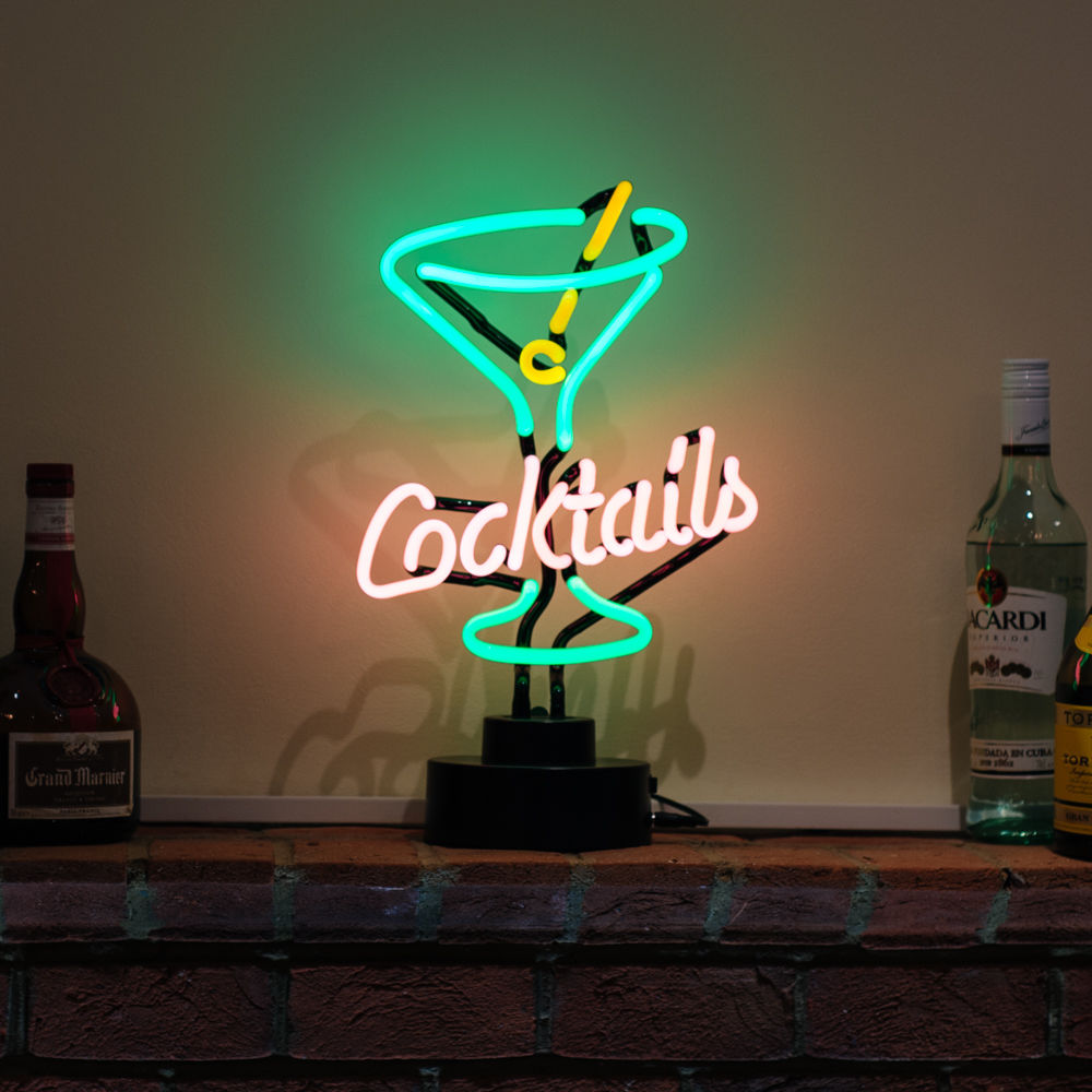 A glowing blue and pink Cocktails glass neon table sign displayed on a wooden surface in front of a home bar setup with various liquor bottles.