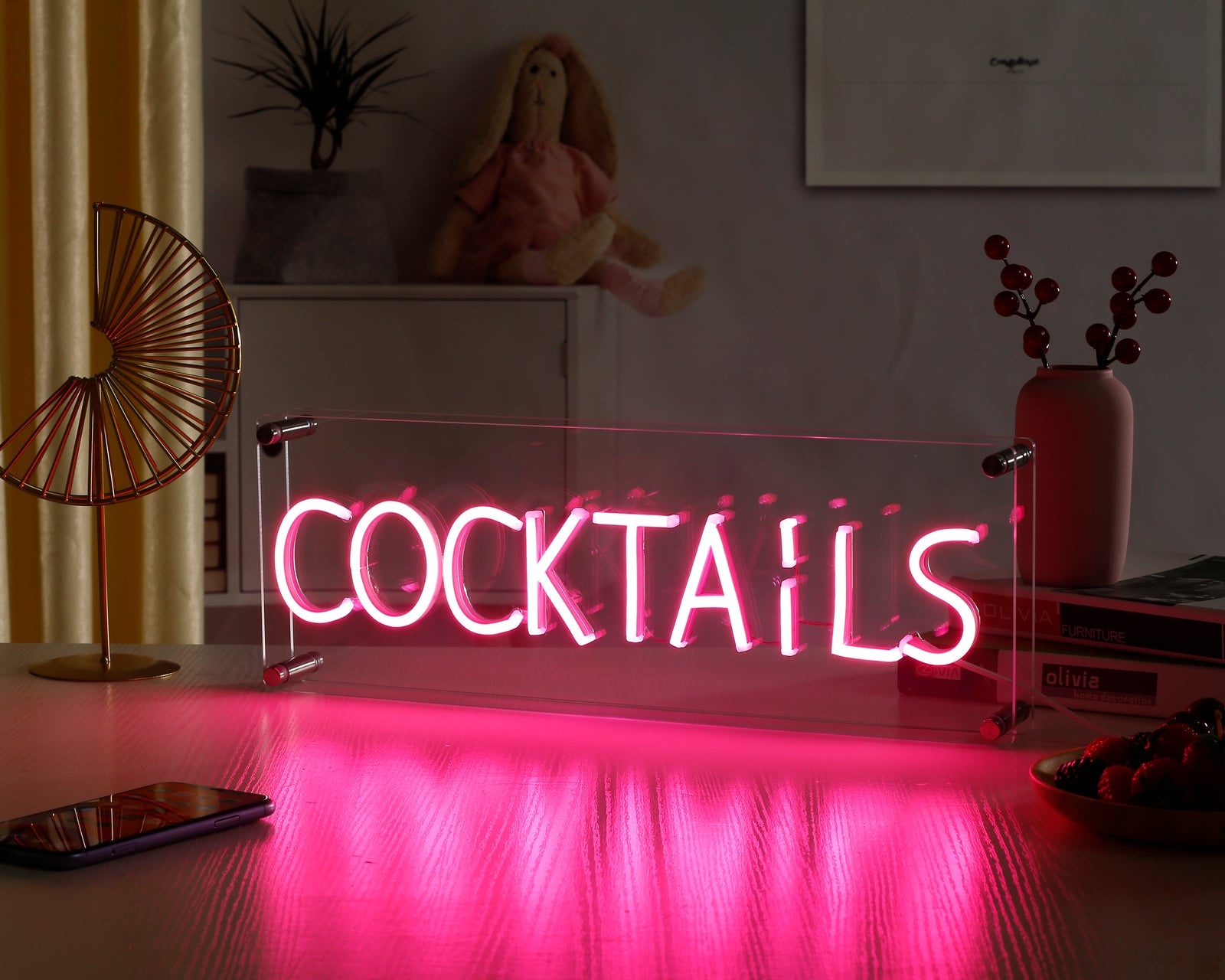 An illuminated "Cocktails" script neon sign glows bright pink and blue inside a clear box on a wooden desk, reflecting slightly below.