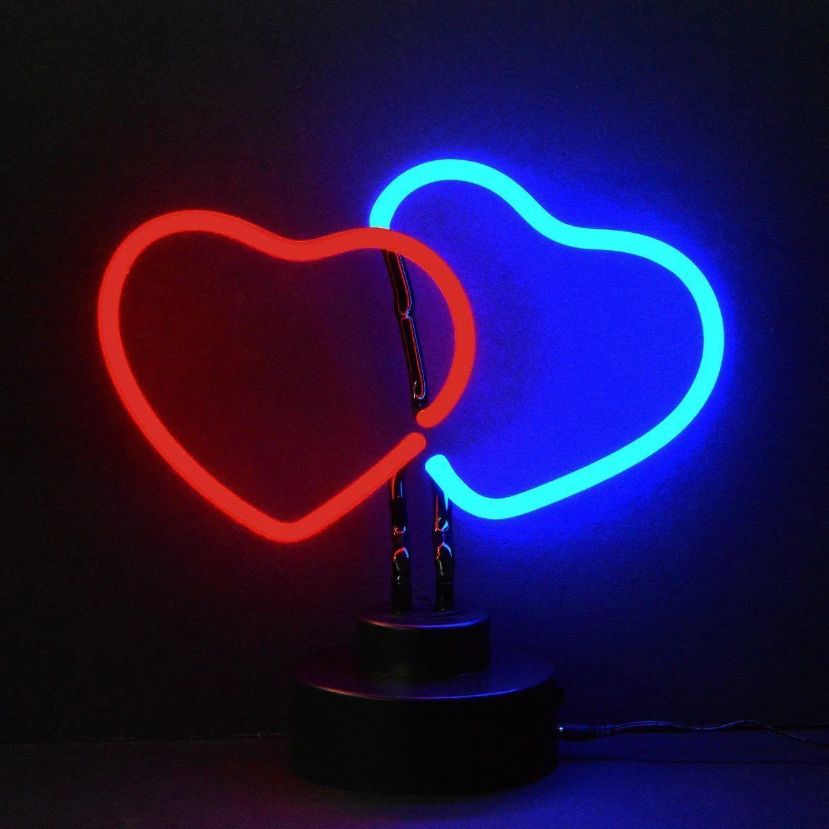 A table-top neon sign displaying two intertwined illuminated hearts glowing orange-red, mounted on a sleek black rectangular base on a wooden desk.