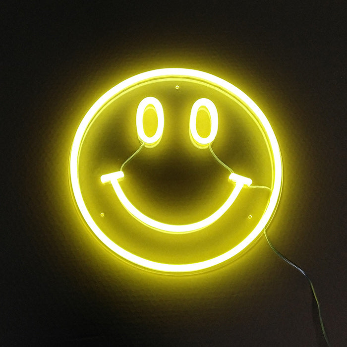 A close-up view of a glowing 'Element Symbol Emoji Smile Face' neon sign mounted on an acrylic backing with a power cord visible.