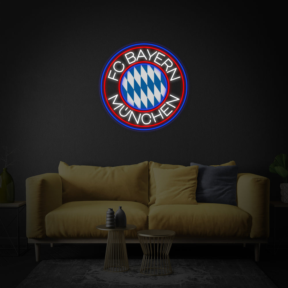 A glowing circular neon sign featuring the FCBM Football design, hanging centrally above a grey sofa in a modern living room setting.