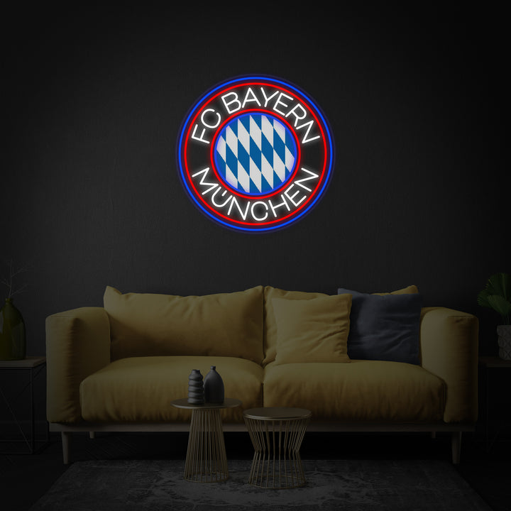 A glowing circular neon sign featuring the FCBM Football design, hanging centrally above a grey sofa in a modern living room setting.