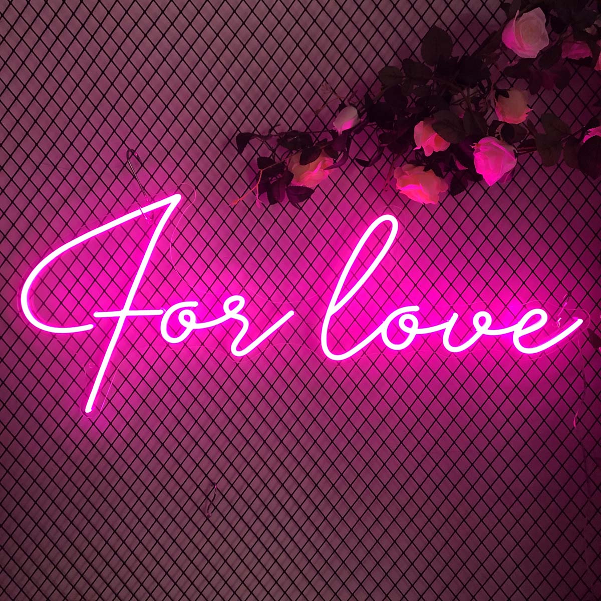 A pink 'For Love' neon wall sign mounted on a wire grid and faux rose floral backdrop.