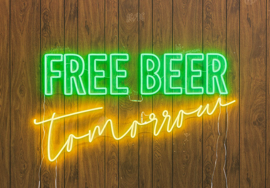 A hand holding a clear acrylic mounted 'Free Beer Tomorrow' neon sign glowing red and blue against a blurred background with doodles, showing the wiring and mounting hardware.