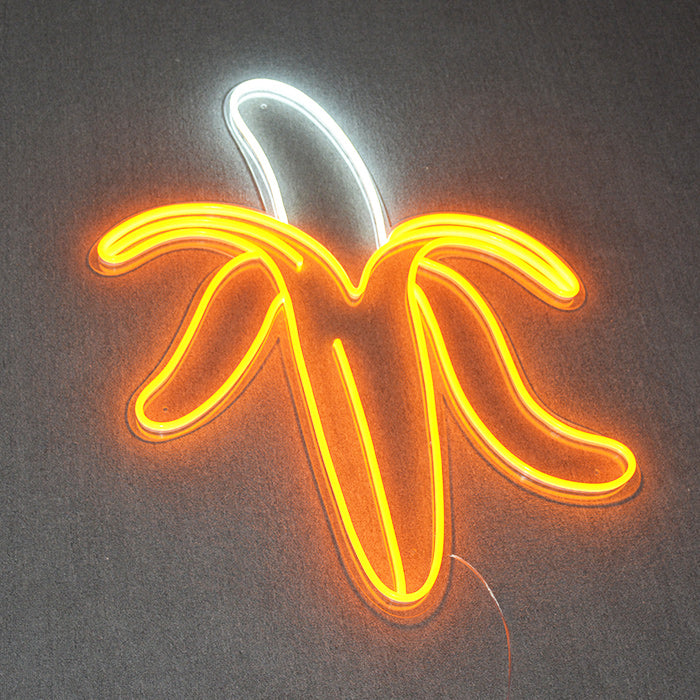 Vibrant yellow glowing neon sign of a banana on a clear backing against a light-colored brick wall