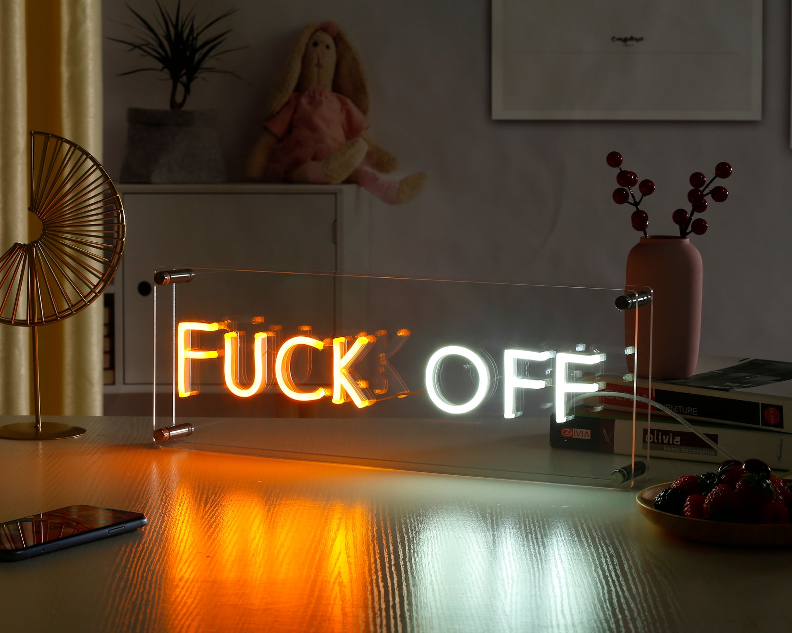 Fuck Off glowing neon sign on an acrylic stand placed on a desk.