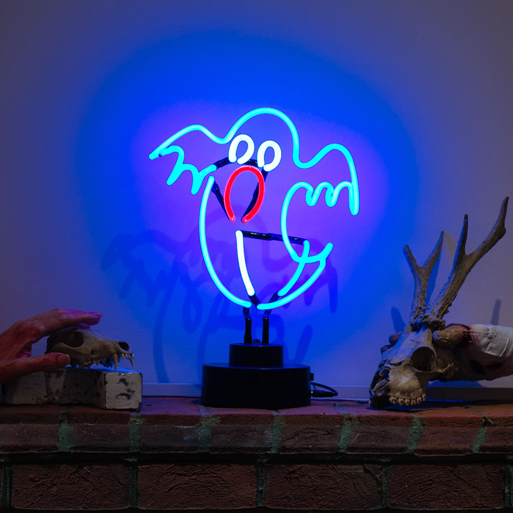 A "Ghost" table neon sign, crafted from real glass tubing, glowing white on a dark wooden surface in a softly lit room, featuring other quirky desk items in the background.