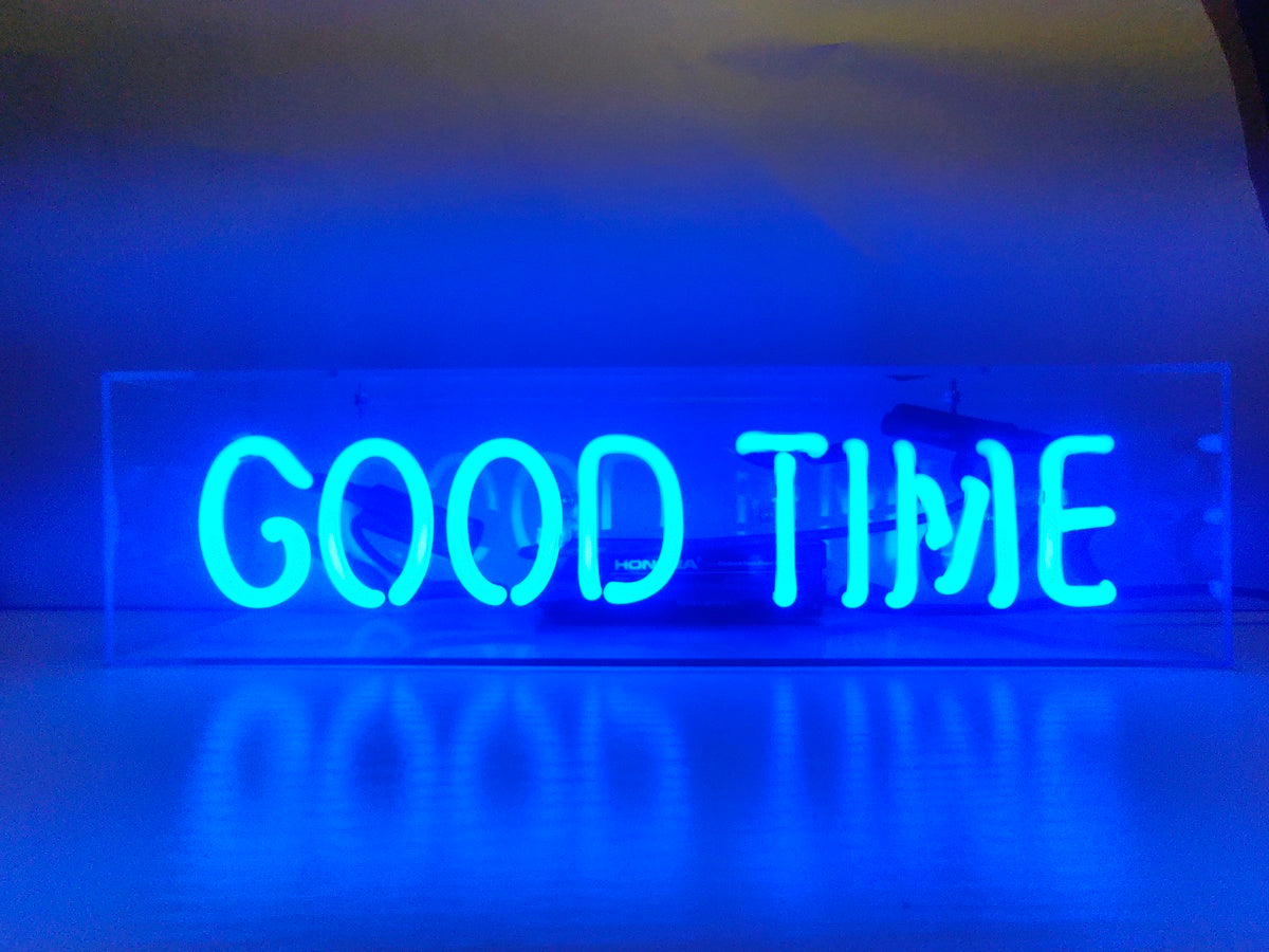 'Good Time' written in a vibrant, glowing red script font within a clear acrylic box neon sign, resting on a reflective wooden surface in a softly lit room.
