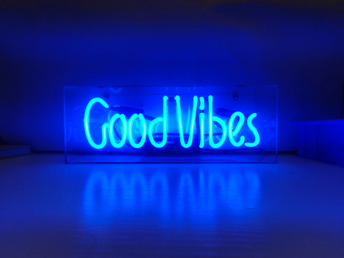 A bright pink handcrafted neon sign spelling "Good Vibes" enclosed in a clear acrylic box and illuminated, sitting on a table with a blurred modern background.