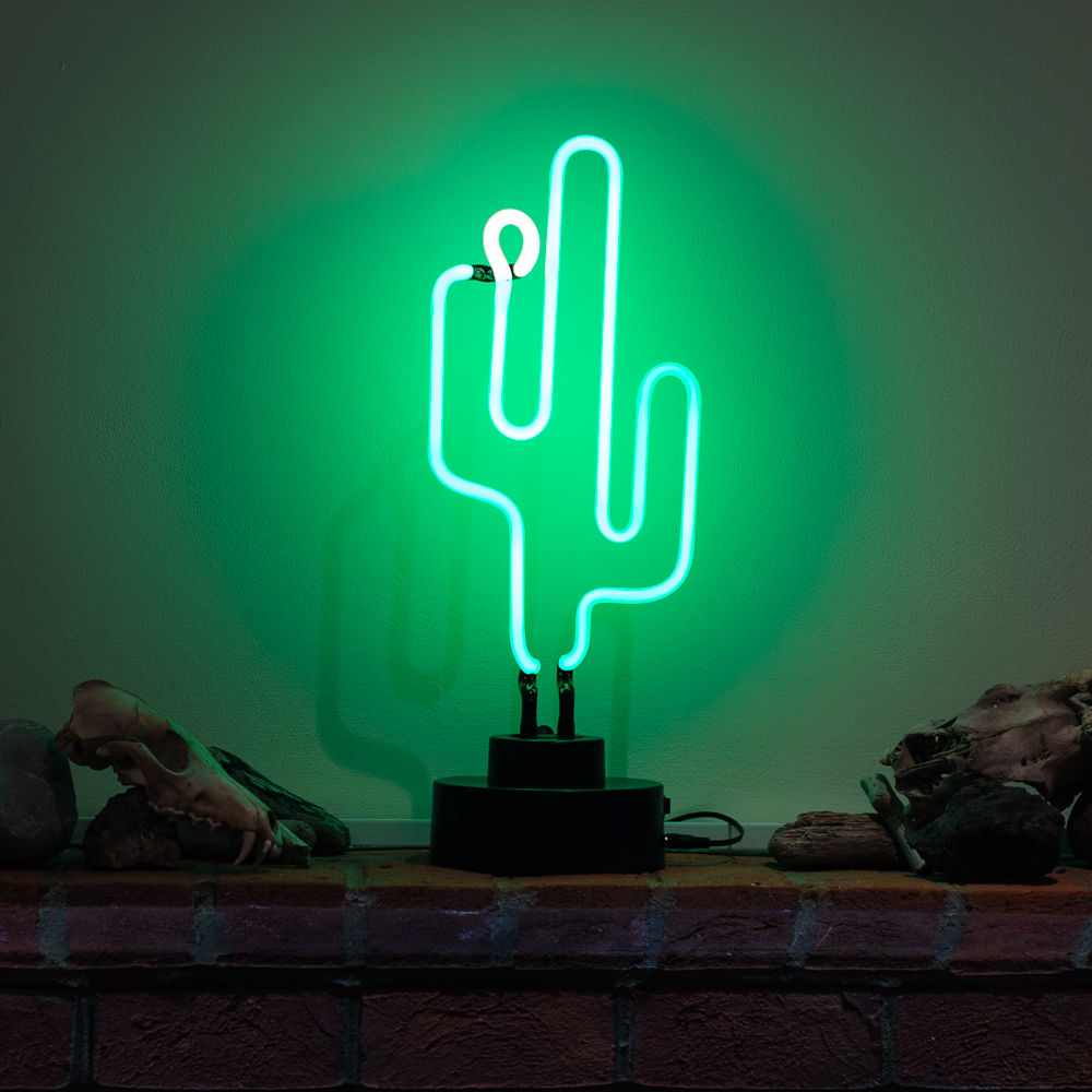 A table neon sign in the shape of a green cactus, featuring green glass tubing lit up on a brick surface next to small decorative skulls and rocks.