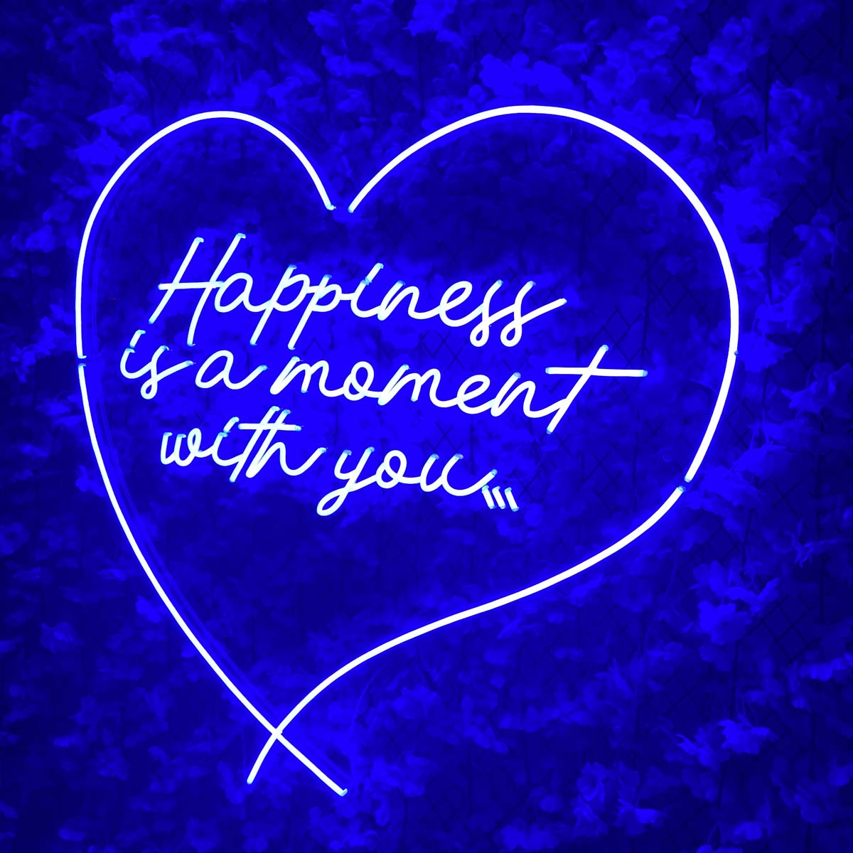 Stylized product image of the glowing text neon sign with 'Happiness is a moment with you' in white cursive, mounted on a stone wall in a cozy room with a plant and book.