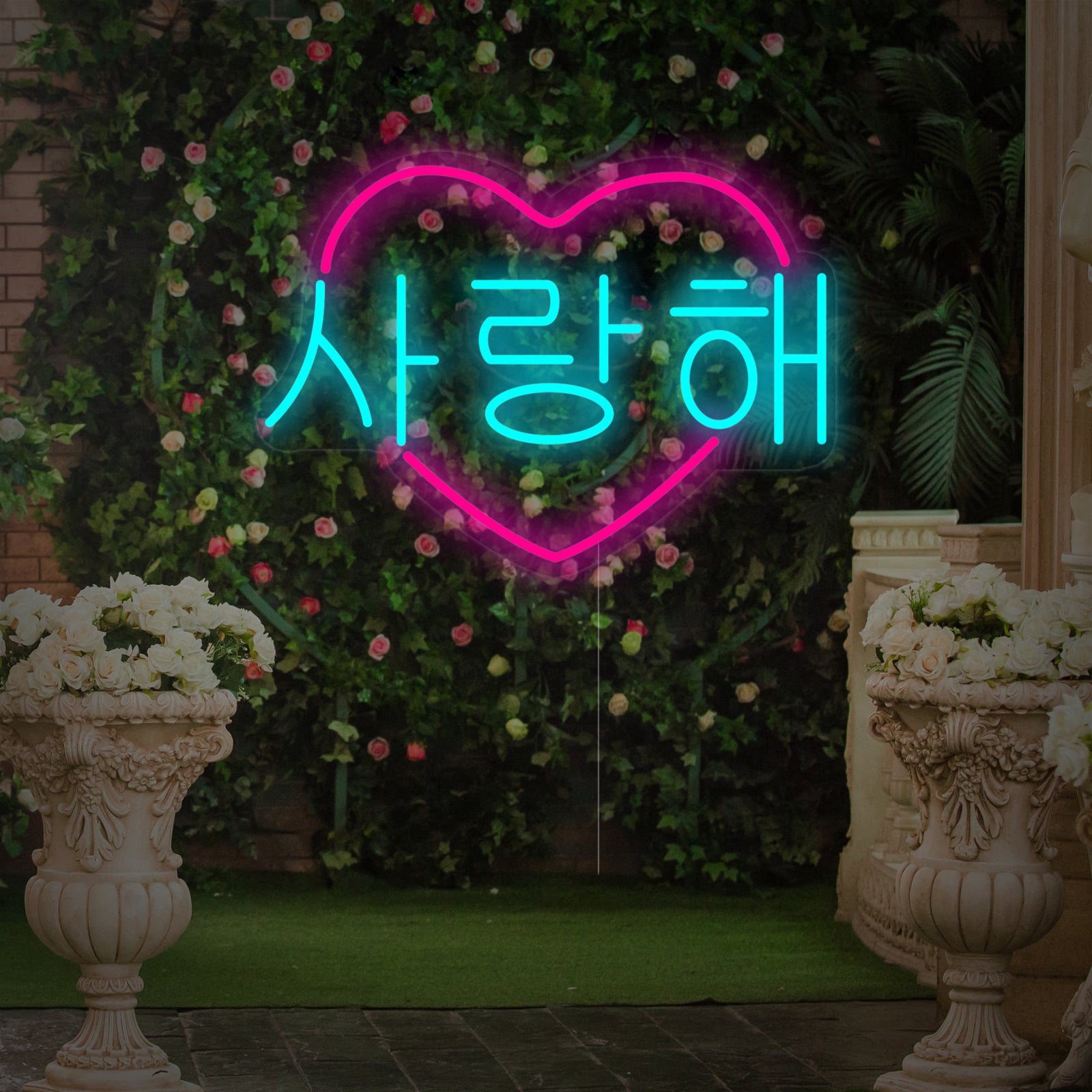 A glowing heart-shaped Korean 'I Love You' neon sign with clear backing, composited against a blurred background of pink flowers.