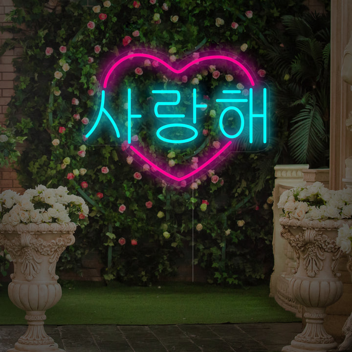 A glowing heart-shaped Korean 'I Love You' neon sign with clear backing, composited against a blurred background of pink flowers.