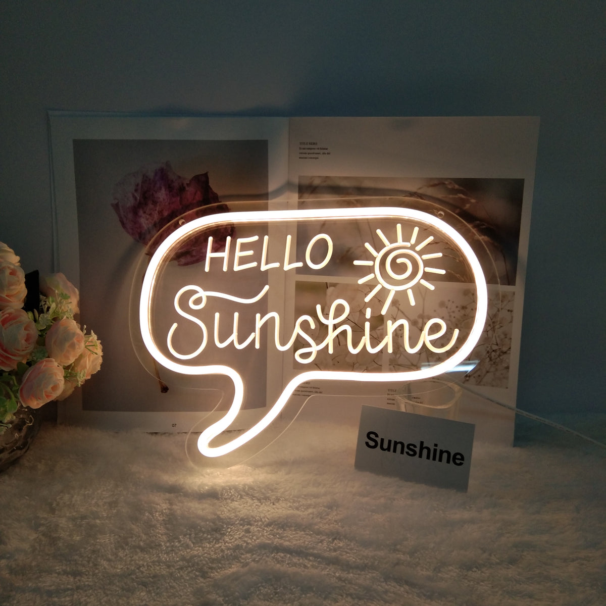 A glowing 'Hello Sunshine' USB mini neon sign on a clear acrylic backing displayed on a desk with books and plants, showing its warm light.
