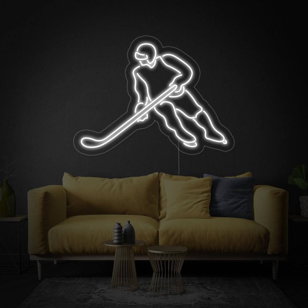 Glowing neon sign of a hockey player in motion, shown hanging on a light grey wall next to a hockey stick and a framed jersey.