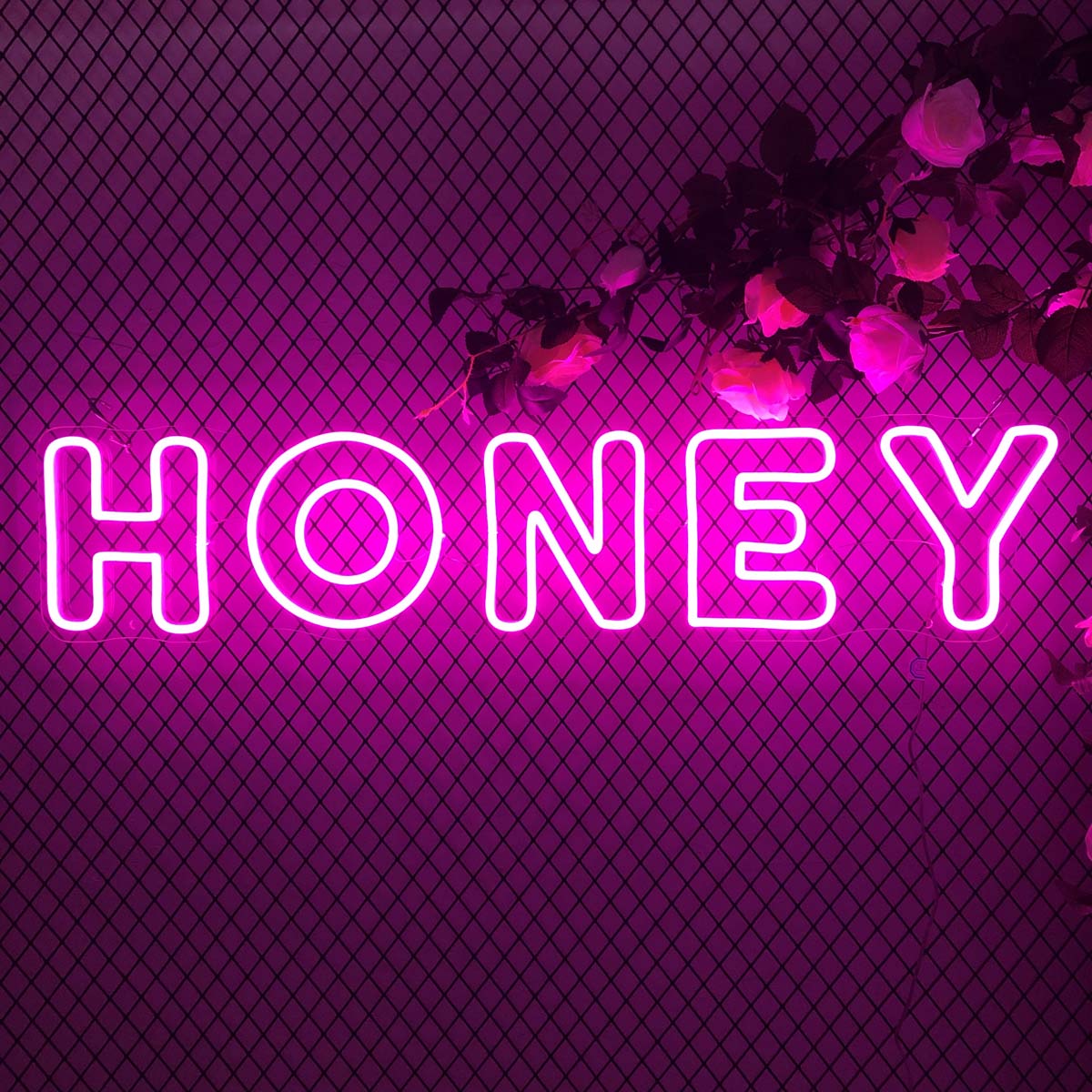 A close-up photograph showing a vibrant, glowing 'HONEY' neon sign rendered in a warm golden-yellow script font, mounted on a rustic black wire mesh grid that's decorated with faux greenery and realistic pink rose blooms.