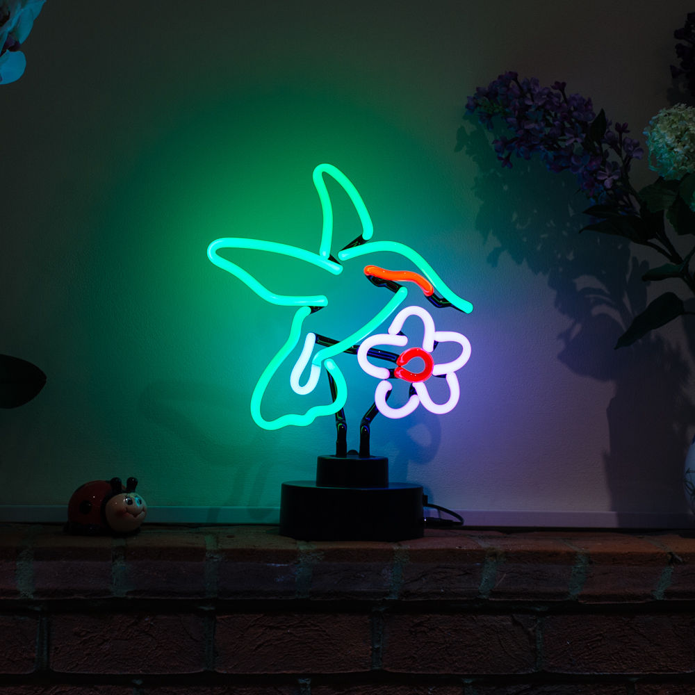 A glowing neon sign of a multi-colored hummingbird perched on a desk next to plants and an open book.