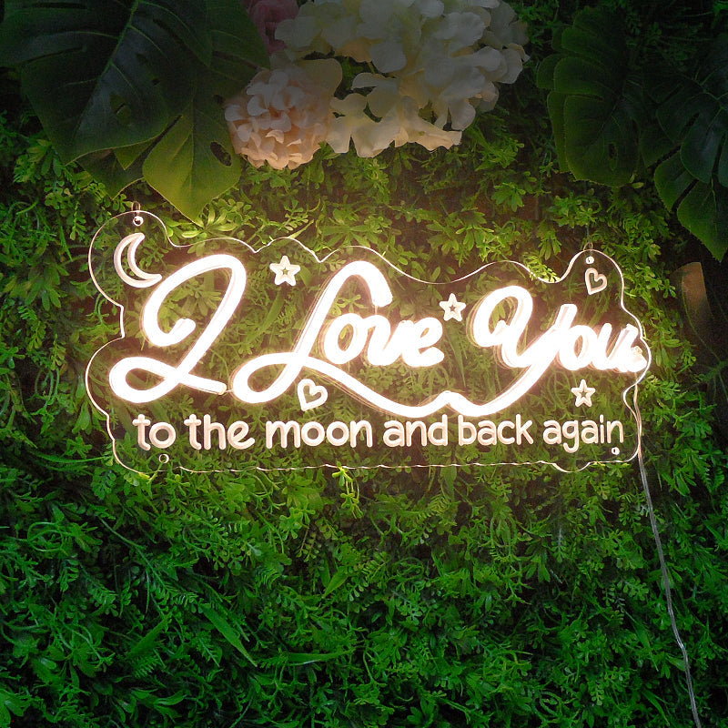 I Love You To The Moon And Back Again neon sign glowing in blue and pink on a green foliage wall above a desk.