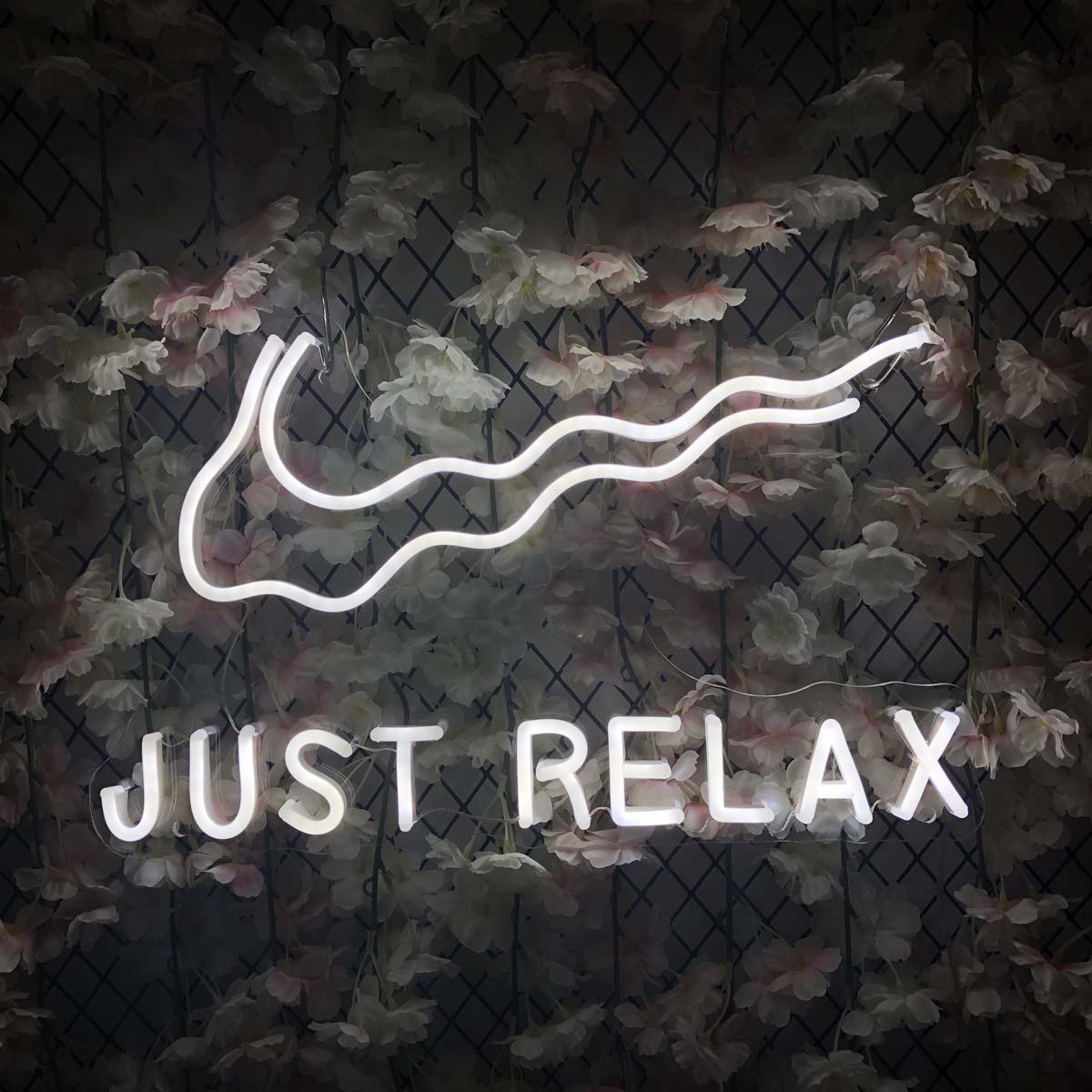 A graphic representation of a 'Just Relax' neon sign featuring elegant script and an illustrative swoosh over a soft background.