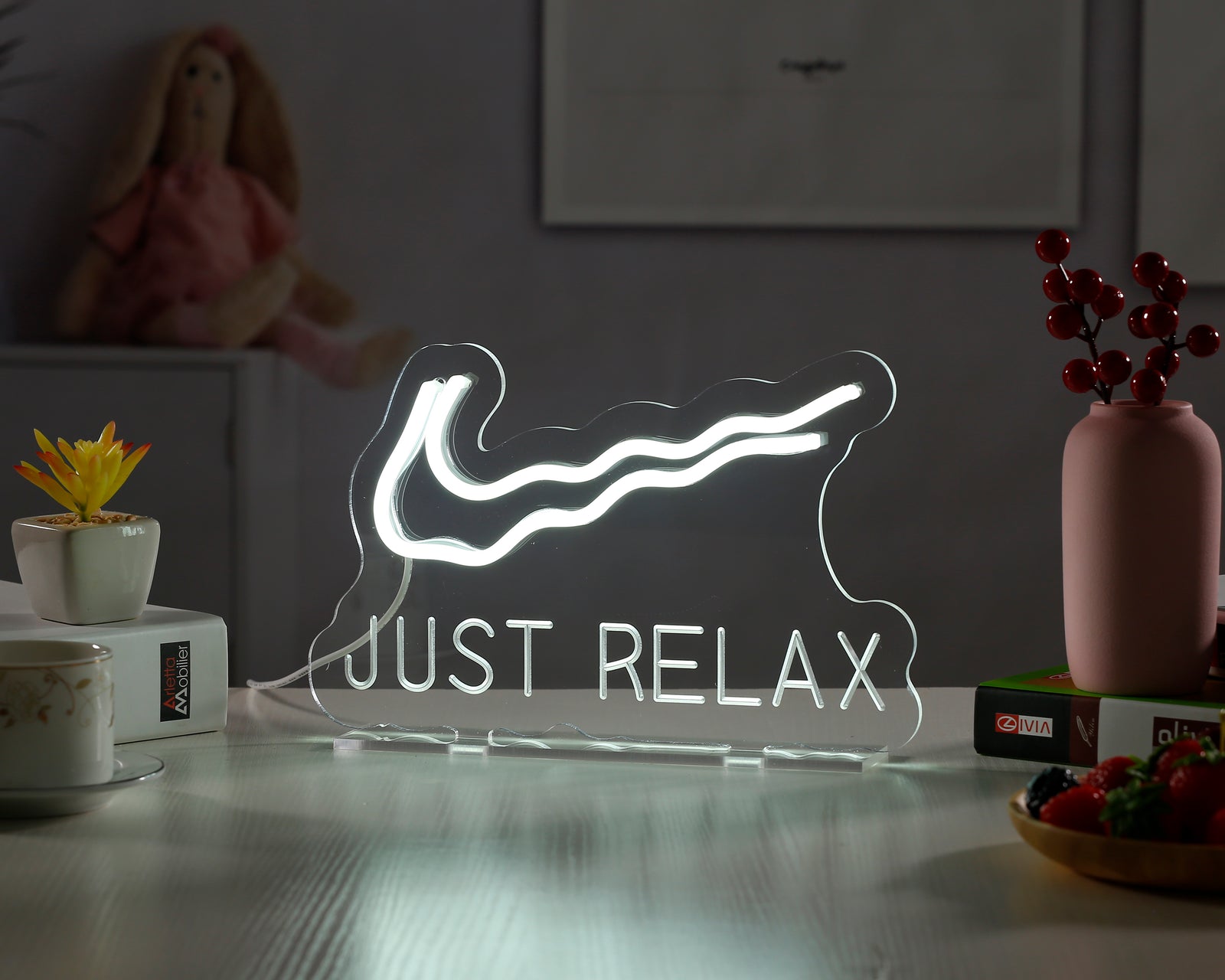 Just Relax glowing white neon desk sign on a wooden surface with potted plant and string lights in background.