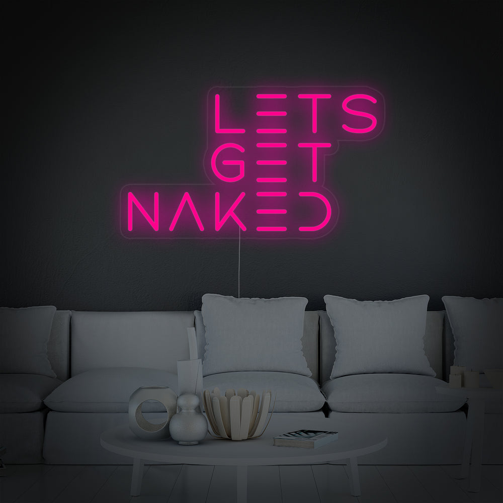 A close-up photograph of the "Let's Get Naked" bathroom neon sign, illuminated in white, clearly showing the script font, acrylic backing, and mounting hardware.