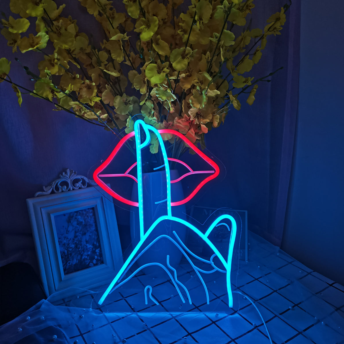 A glowing red and white neon sign on an acrylic backer depicting red lips and the words 'Be Quiet' positioned against a soft-focus background with plants and light reflections.