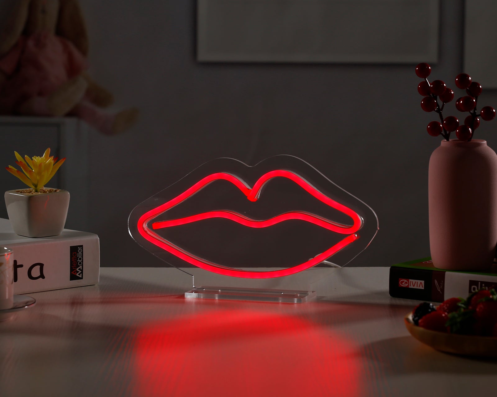 A clear, close-up photograph of a red neon sign shaped like a pairs of lips glowing vividly while sitting on its clear acrylic desk stand, set against a blurred background with potted plants and a woven basket.
