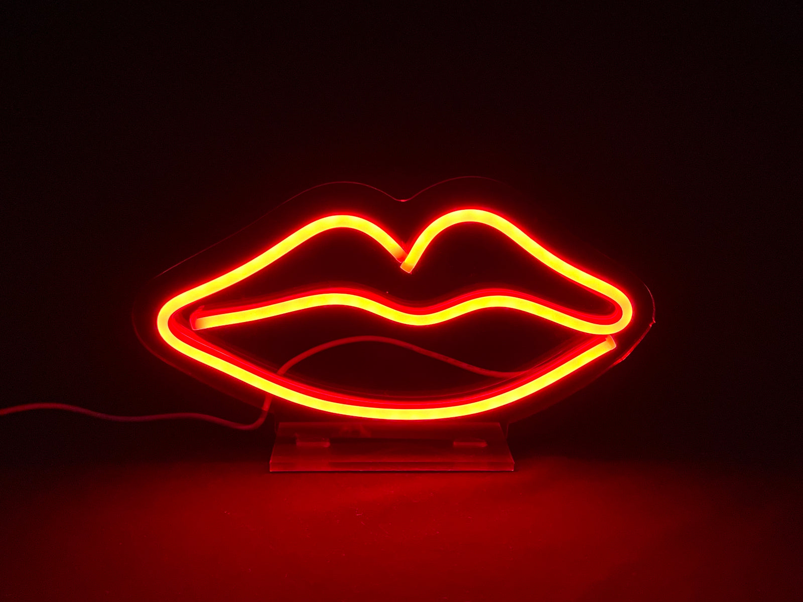 A front-on photograph of a glowing red neon sign shaped like lips with a matching red outline, resting on a flat, reflective dark surface that mirrors the red light and the clear acrylic base.