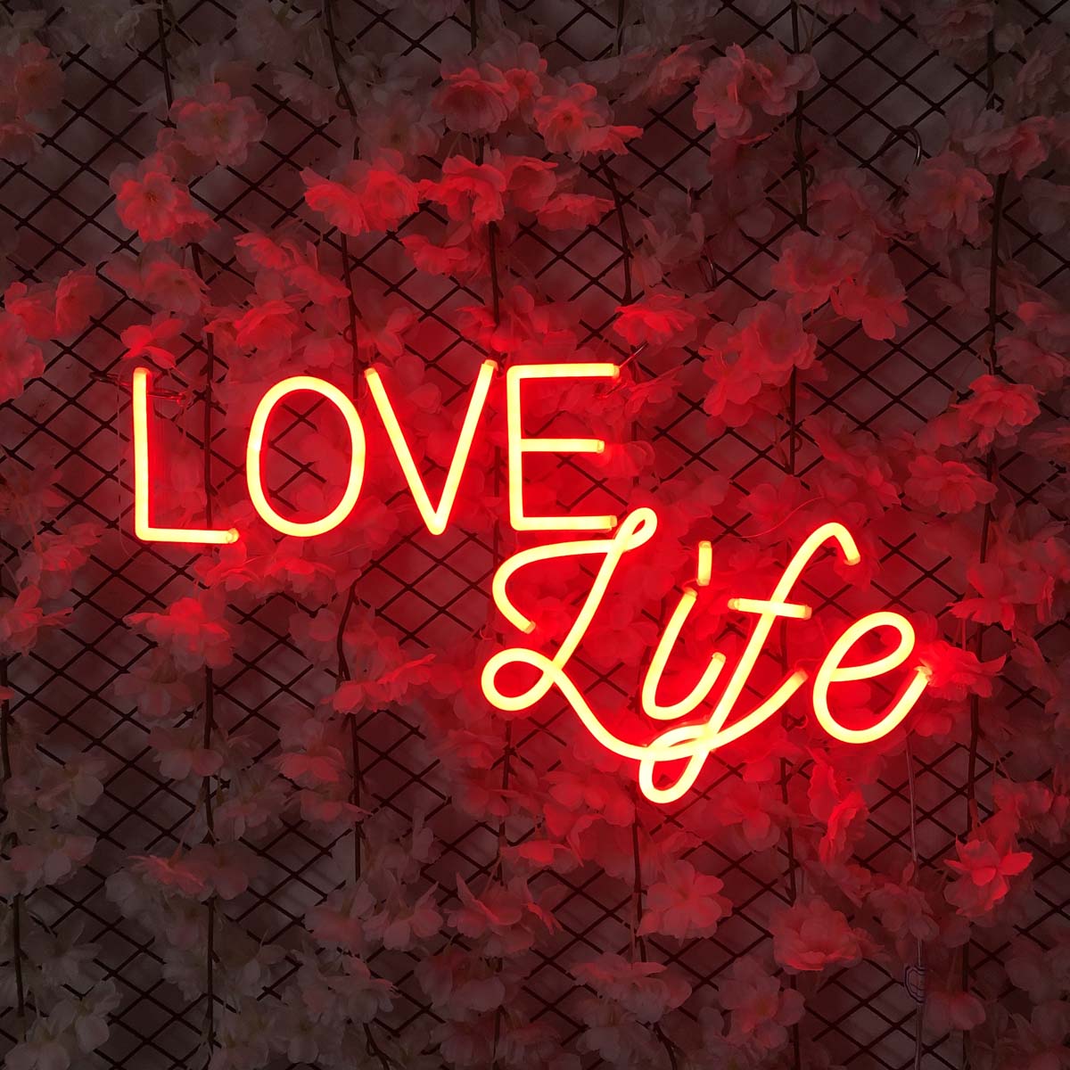 A illuminated 'Love Life' neon wall sign in a modern script font, mounted on a wire grid panel among a decorative arrangement of artificial flowers and foliage.