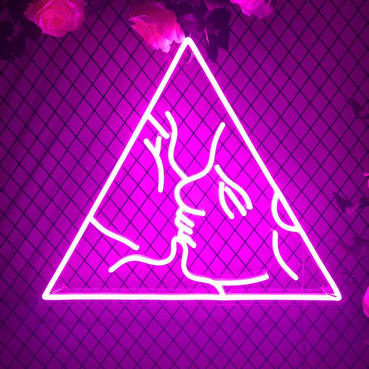 A glowing neon sign depicting a man and a woman in a close embrace and kiss, glowing warm yellow against a brick wall accented with climbing roses.