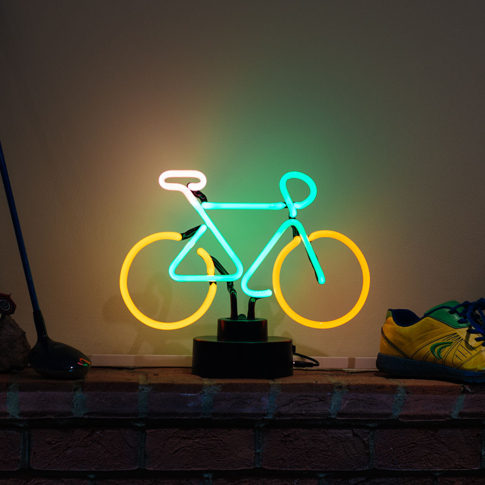A glowing blue and orange glass mountain bike table neon sign on a clear acrylic stand, positioned against a rustic brick wall, showing the power cord connection.