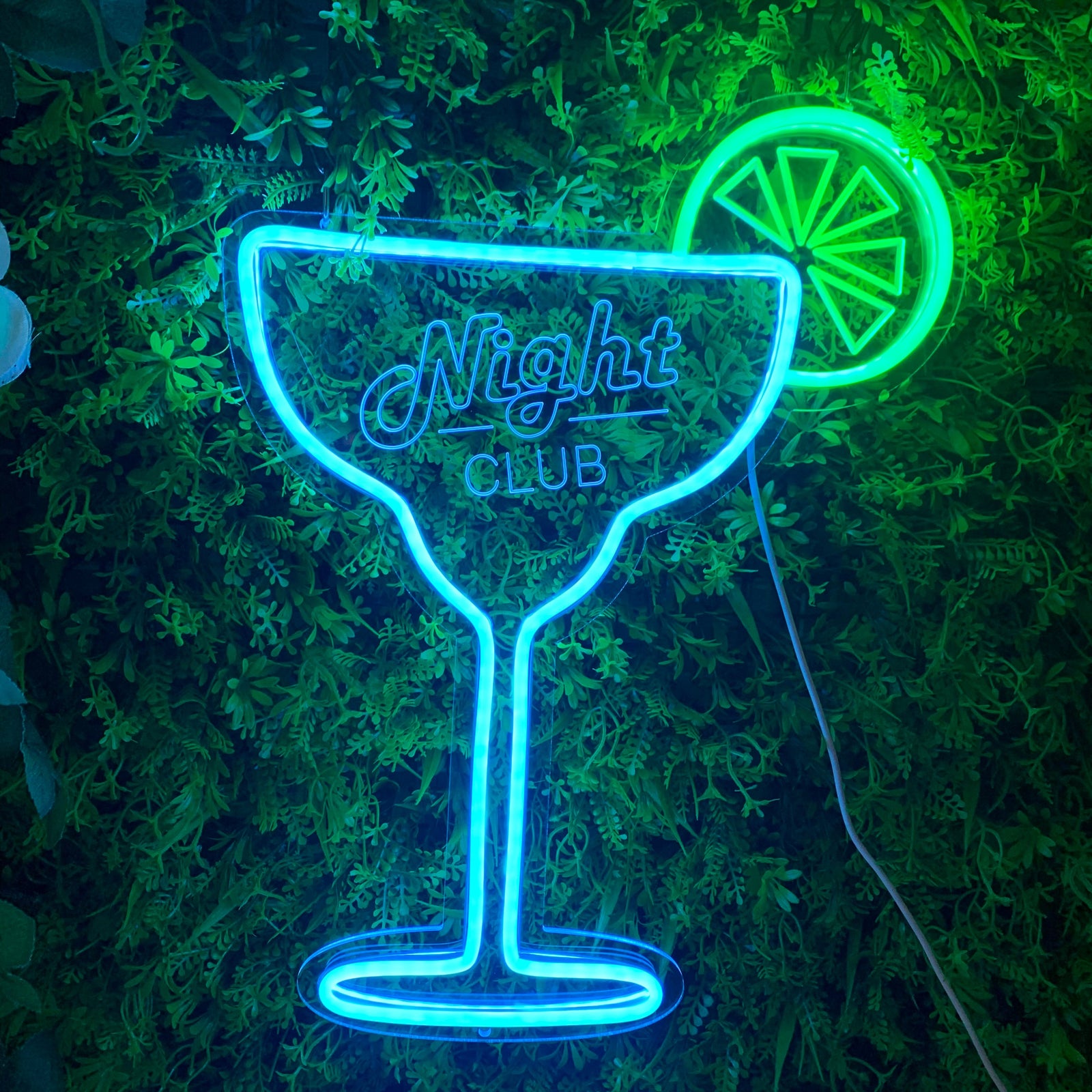 A illuminated blue and pink cocktail glass mini neon sign against a dark artificial foliage background