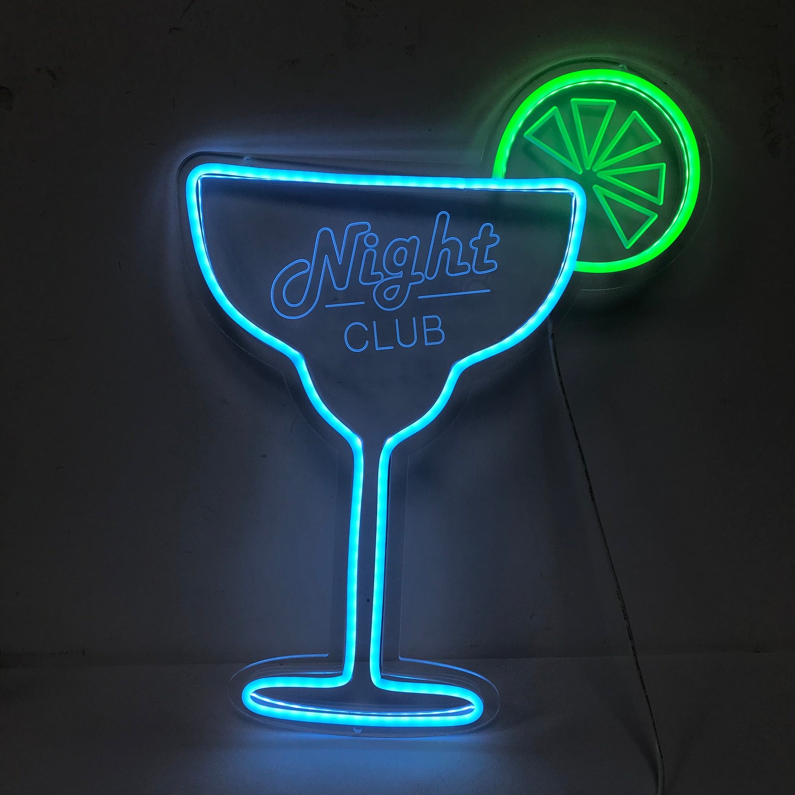 A close-up photograph of an illuminated cocktail glass mini neon sign hanging on a dark artificial leafy wall