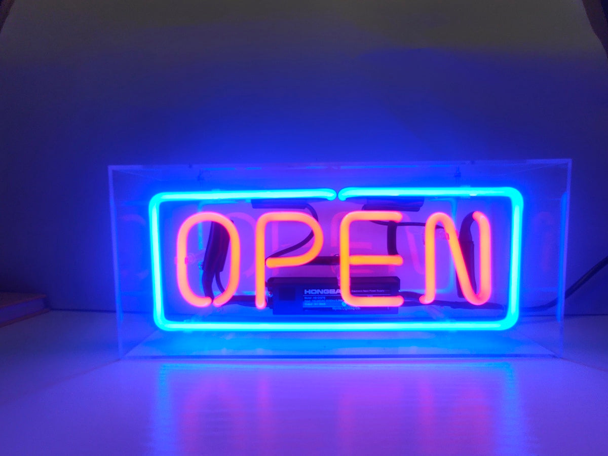Handcrafted neon sign featuring glowing red 'Open' text and a teal border, encased in a clear acrylic box, shown sitting on a shelf with internal transformer visible.