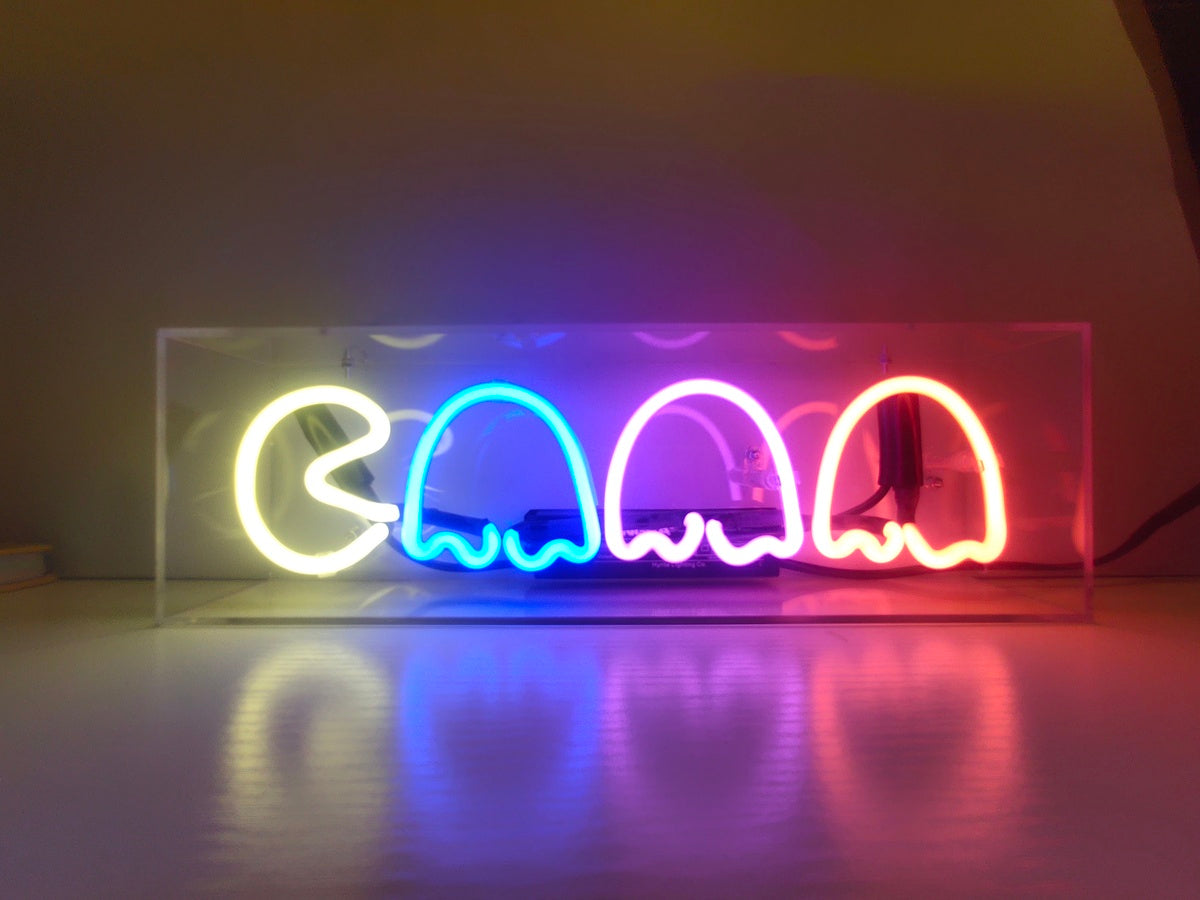 Pac Ghost blue neon sign glowing inside a clear acrylic display box on a dark wooden table in a dimly lit room, with visible internal wiring and reflections.