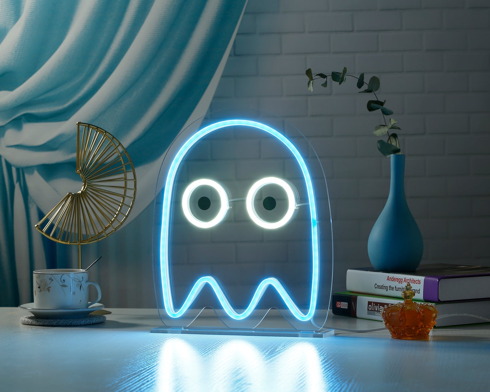 A glowing blue ghost desk neon sign illuminated in a dimly lit room, featuring a clear acrylic backing and a dark stand, with a curtain and plant blurred in the background.