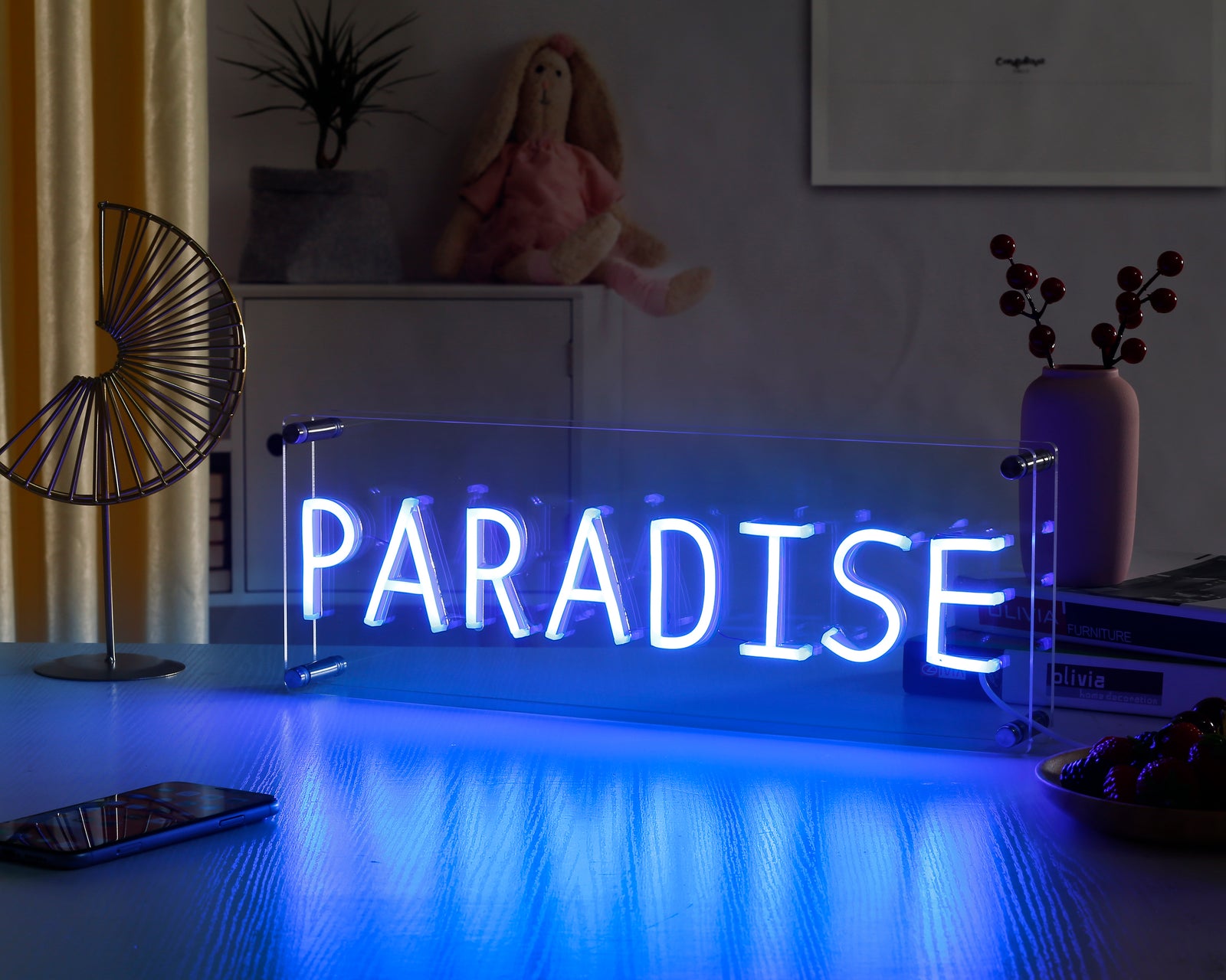 A close-up photograph of a pink Paradise desk neon sign, illuminated on a table in a dimly lit room with blurred background lights.