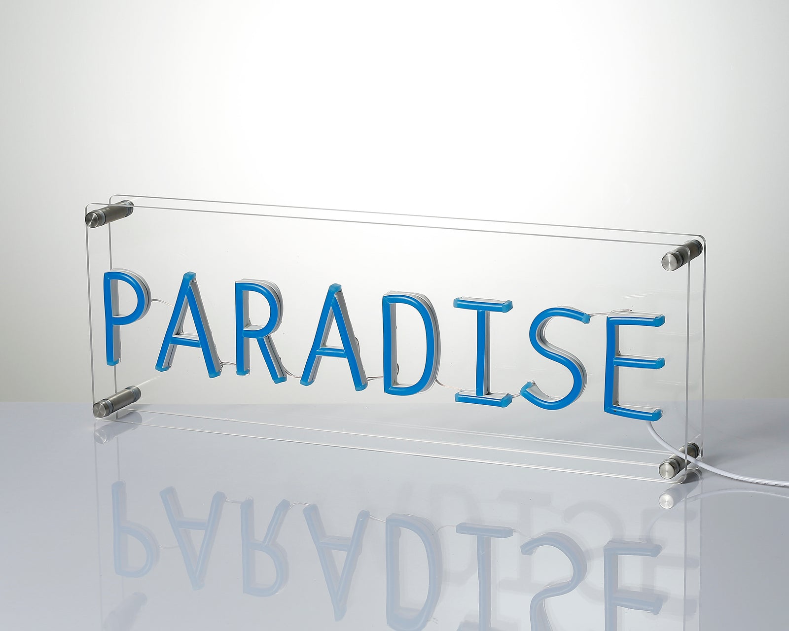 A glowing pink Paradise neon sign in a scripted font, sitting on a wooden desk next to a plant and other small decor items.